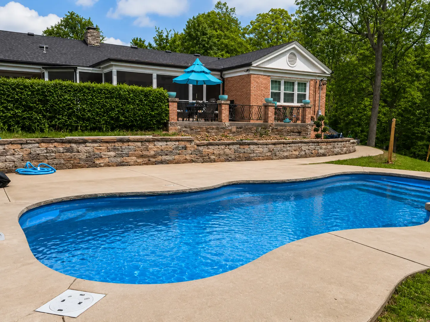 Enhanced freeform fiberglass pool with blue water, brick home, raised patio, and landscaped retaining wall in Columbia, IL.